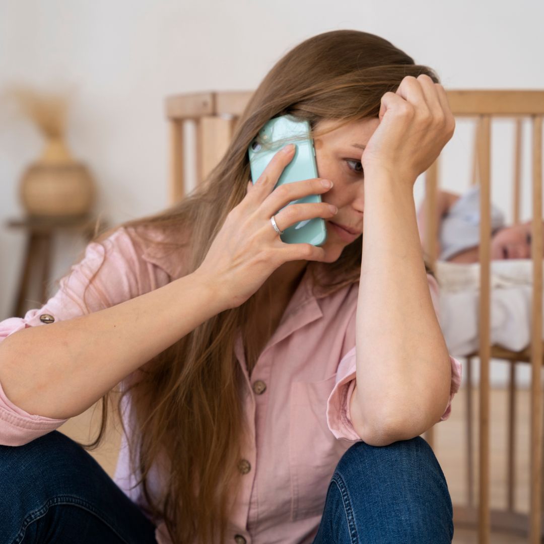 Woman in pink shirt on phone, concerned, with crib nearby; postpartum insomnia