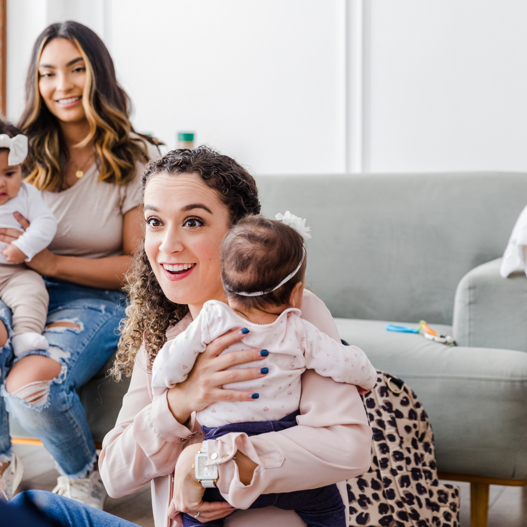 Woman in pink blouse holding baby wearing pink bow; couch with leopard pillow nearby