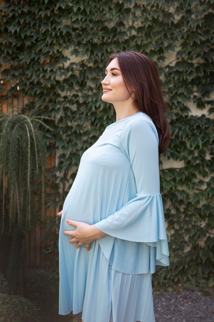 Pregnant woman in light blue ruffled dress in a lush garden, showcasing maternity fashion