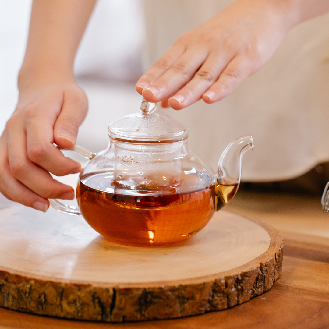 Hands handling teapot with amber tea on rustic wood, smartphone not visible.