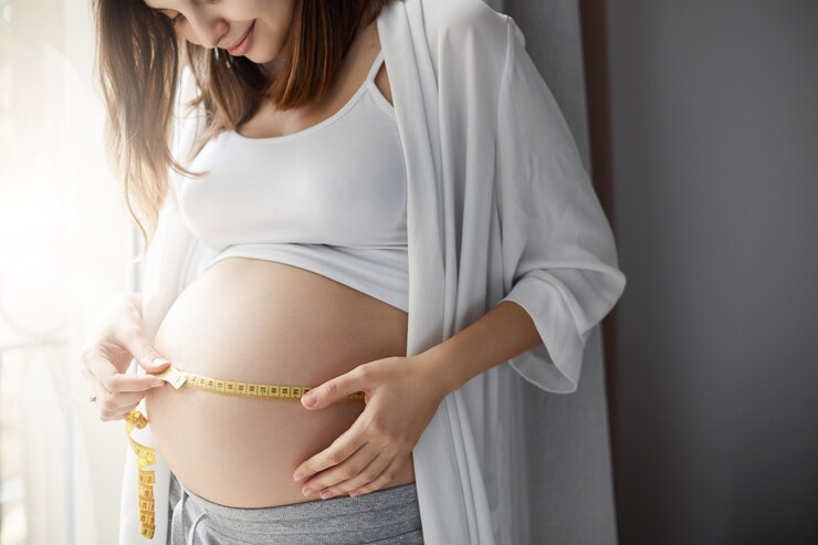 Pregnant woman with measuring tape, wearing robe and tank top, in softly lit room