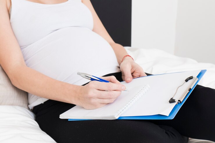 Woman writing in notebook on bed, focus on Mommy Makeover contemplation