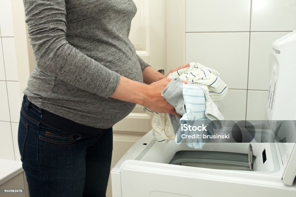 Person loading washing machine with laundry in gray shirt and blue maternity pants.